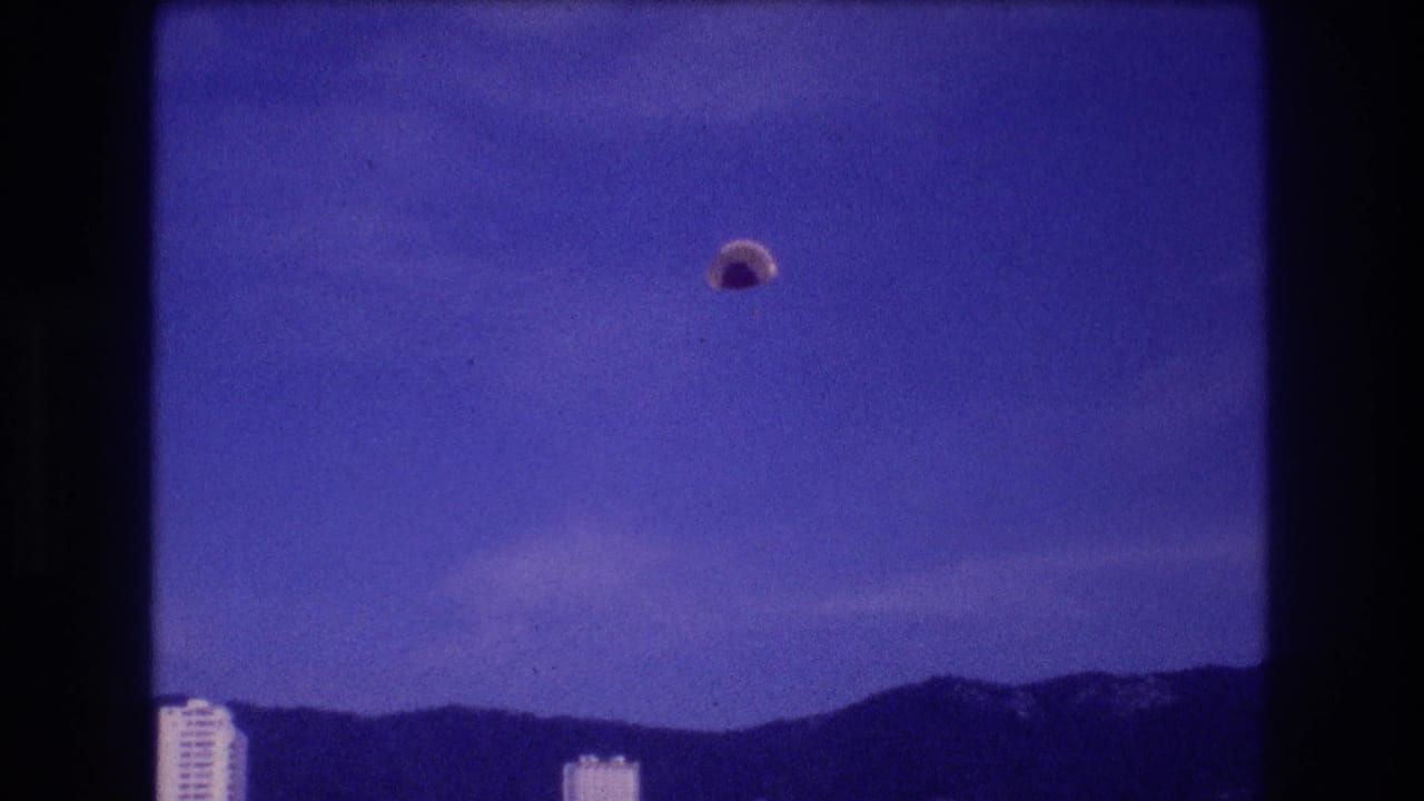 8mm home-movie frame, Acapulco c. 1982: parachute canopy descended lower, rooftops of high-rise hotels at bottom edge, mountain range behind.