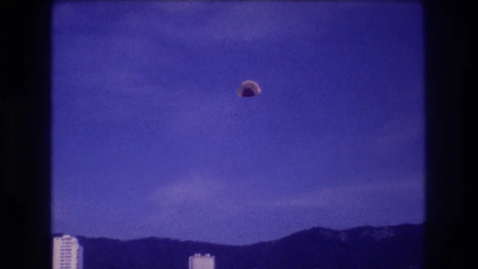 8mm home-movie frame, Acapulco c. 1982: parachute canopy descended lower, rooftops of high-rise hotels at bottom edge, mountain range behind.
