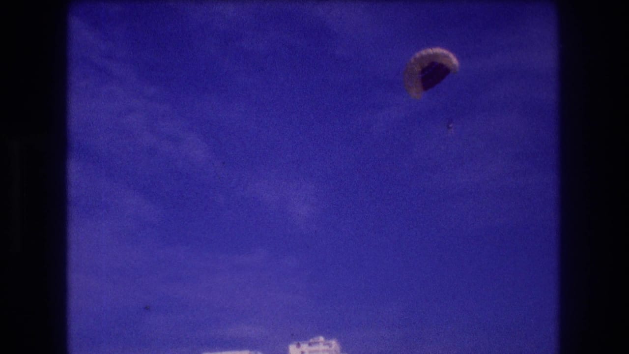 8mm home-movie frame, Acapulco c. 1982: parachute canopy large in frame near end of descent, hotel rooftops at bottom edge, faint mountain outline.