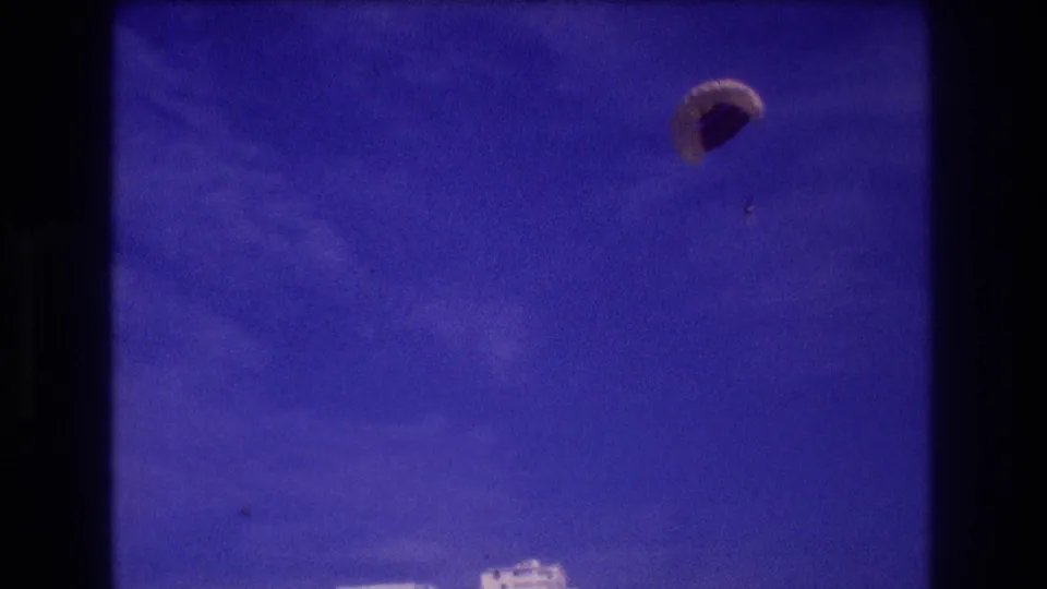 8mm home-movie frame, Acapulco c. 1982: parachute canopy large in frame near end of descent, hotel rooftops at bottom edge, faint mountain outline.