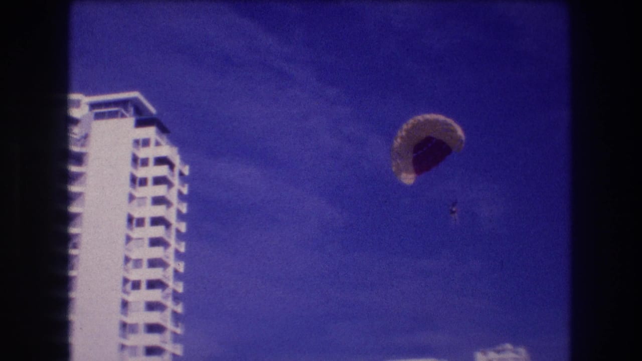 8mm frame from Acapulco, 1982 — parasailer descends past white resort hotel building as palm-tree tops enter the bottom of the handheld shot.