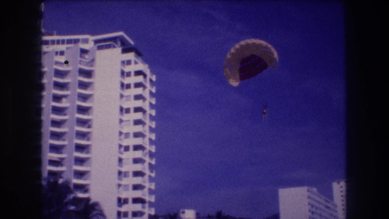 Final 8mm frame from Acapulco, 1982 — parasail canopy has drifted rightward; two white resort buildings and palm foliage now frame the shot.