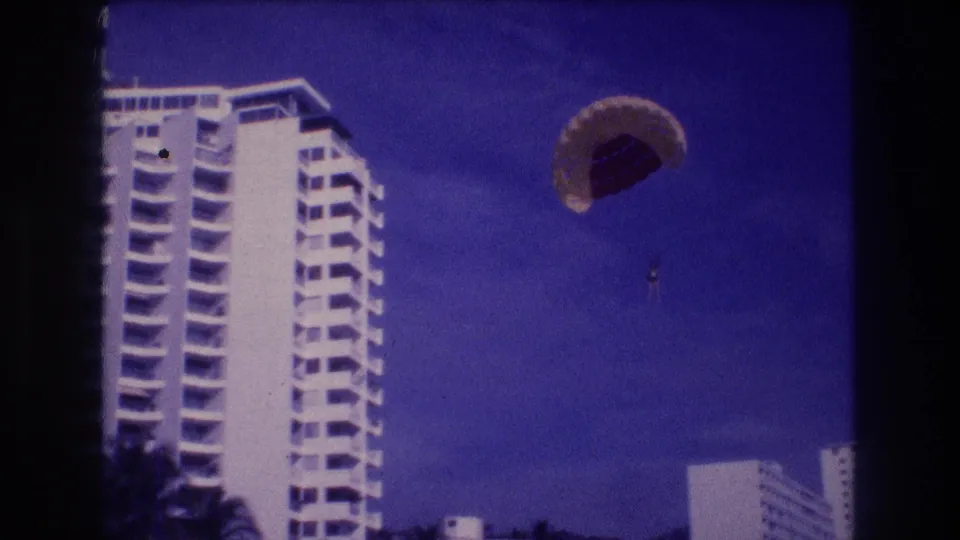 Final 8mm frame from Acapulco, 1982 — parasail canopy has drifted rightward; two white resort buildings and palm foliage now frame the shot.