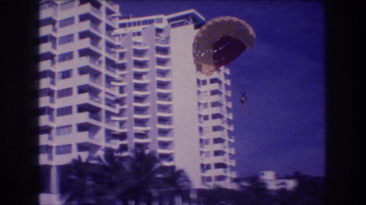 Parasail canopy larger and lower as it descends beside the same high-rise resort hotel, palm trees in foreground