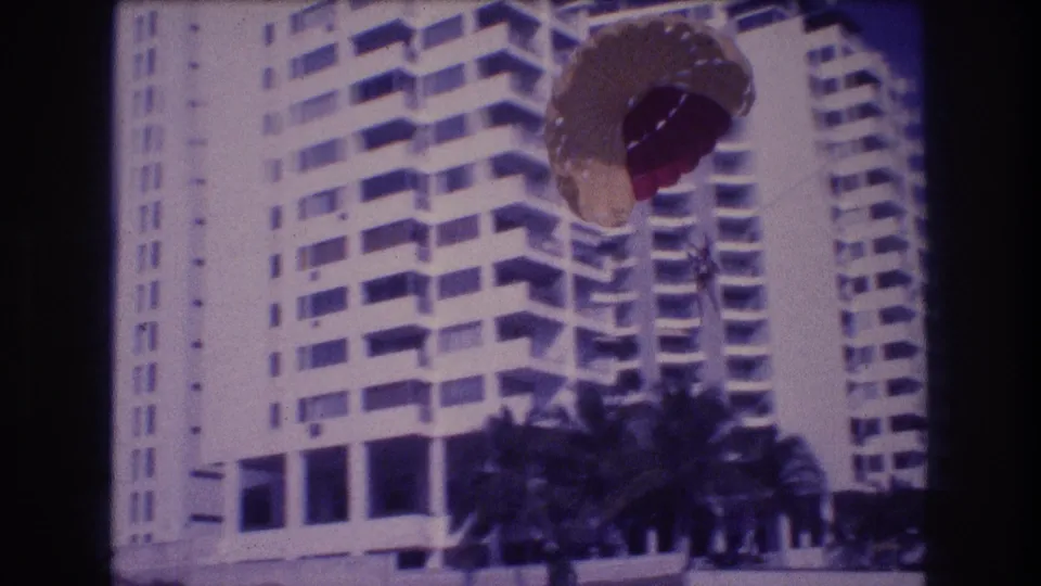 Super 8 home movie, c.1982 Acapulco: close view of parasail canopy with resort hotel and palm trees as parasailor nears the beach