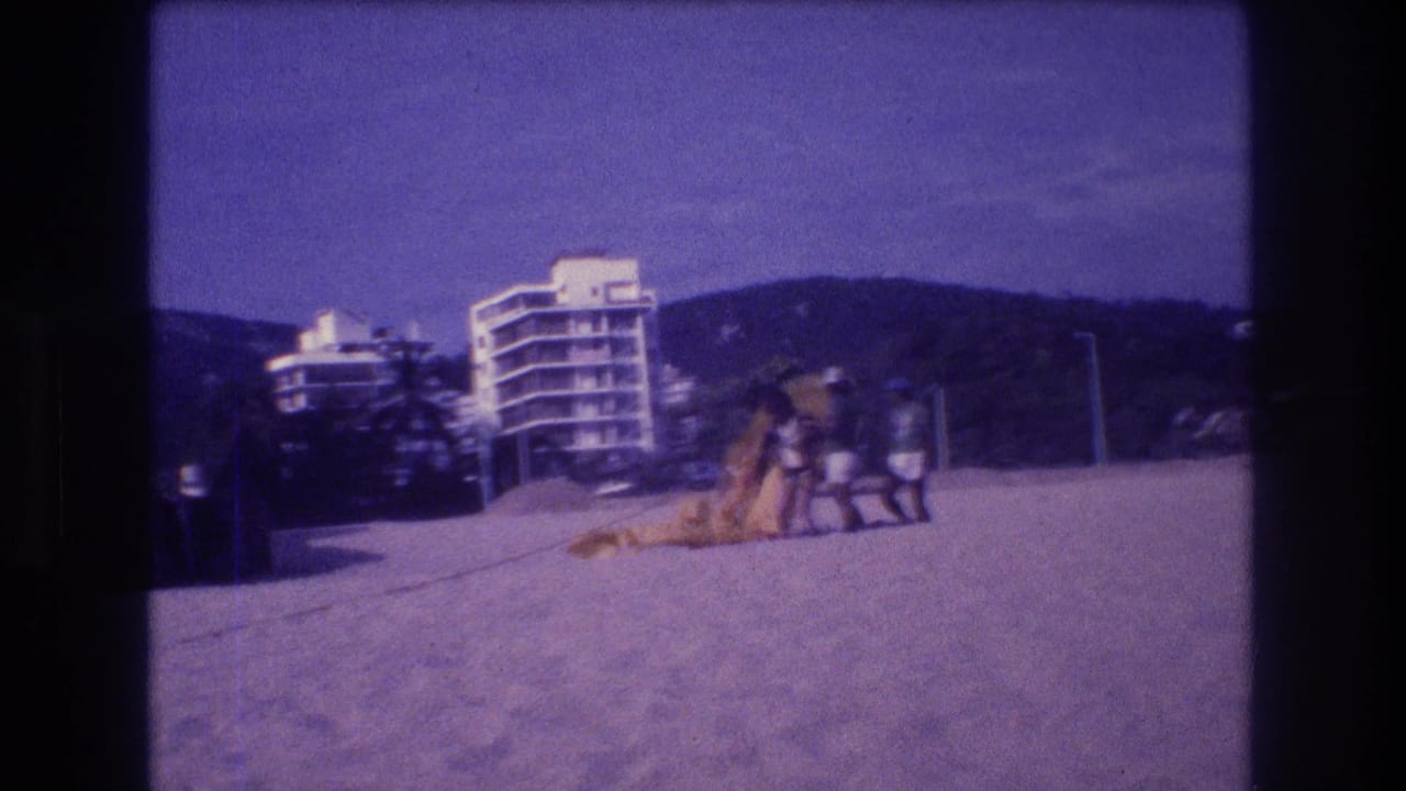 1982 Acapulco beach on 8mm film: figures stand near a deployed parachute on open sand, with a modernist hotel building and palm-covered hillside visible.