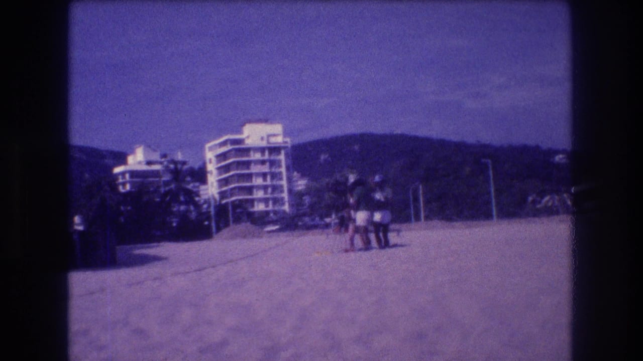 Wide beach shot with a small group on open sand; multi-story hotel, palm trees, and hillside visible in background, parachute less prominent.