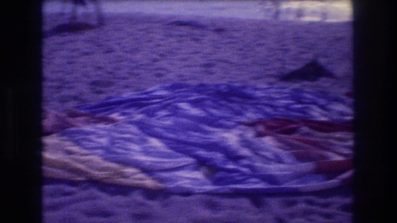 Colorful patterned beach towel spread on purple-tinted sand with a person partially reclining on it.