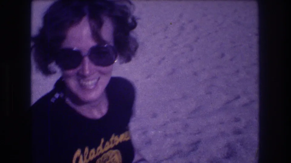 Smiling woman in oversized round sunglasses and dark graphic t-shirt on sandy beach; 8mm home movie, Acapulco, Mexico, 1982, with purple color shift from aged film stock.