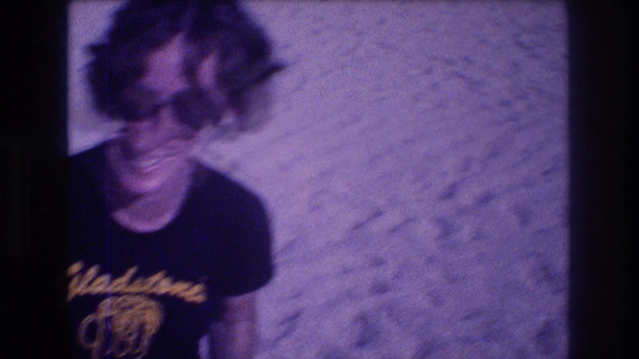 Woman with curly hair and large dark sunglasses laughing, black t-shirt with yellow Gladstone's script logo, sandy beach background, heavy purple color cast.