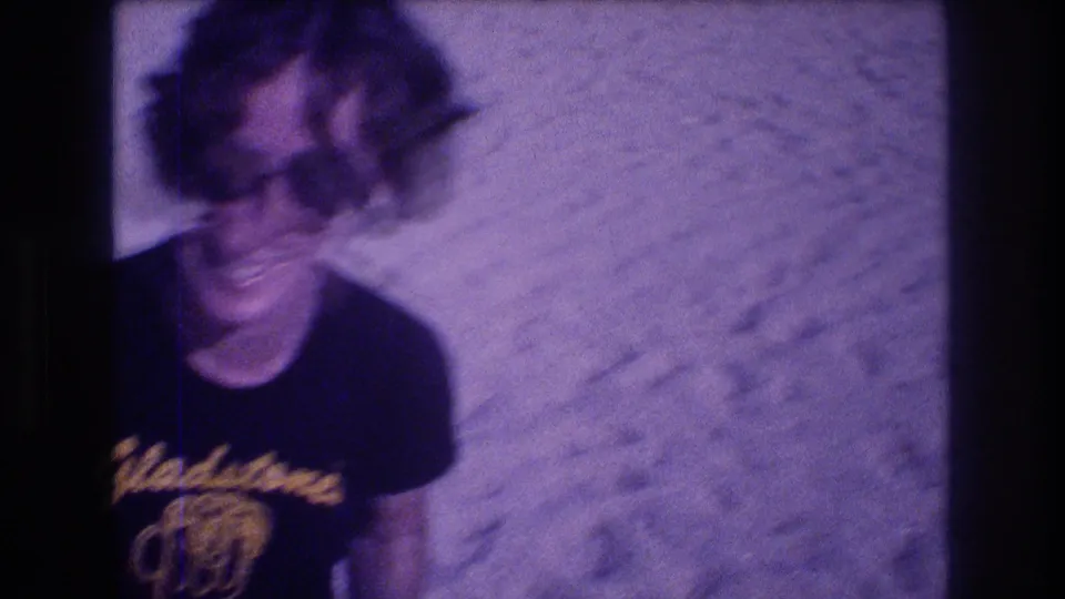 Woman laughing toward camera on a beach, metadata-dated Acapulco, Mexico, 1982; black souvenir t-shirt with Gladstone's logo, oversized sunglasses, heavy 8mm purple fading.