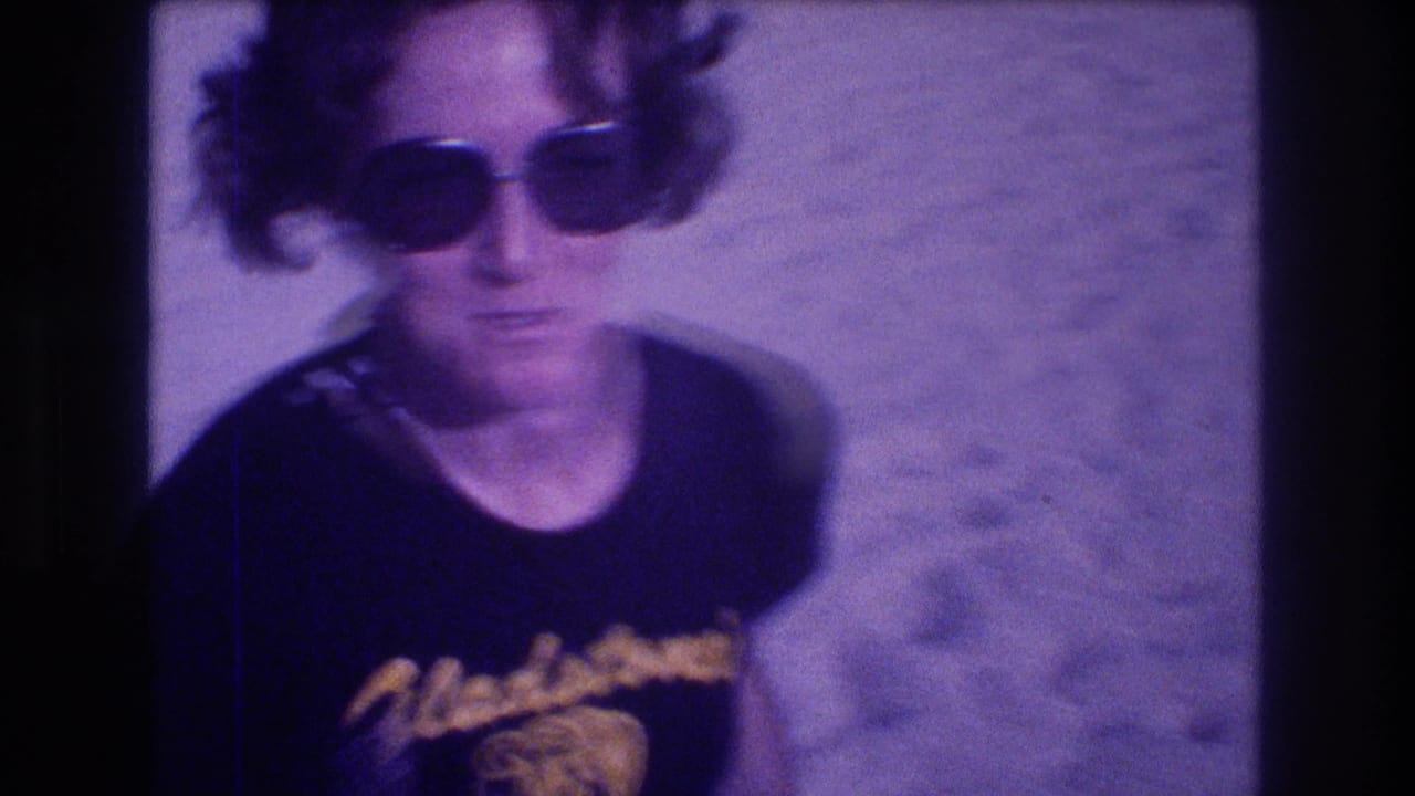 Close portrait of a woman in oversized dark sunglasses and black Gladstone's souvenir shirt on a sandy beach; metadata-dated Acapulco, Mexico, 1982, 8mm film with pronounced purple shift.