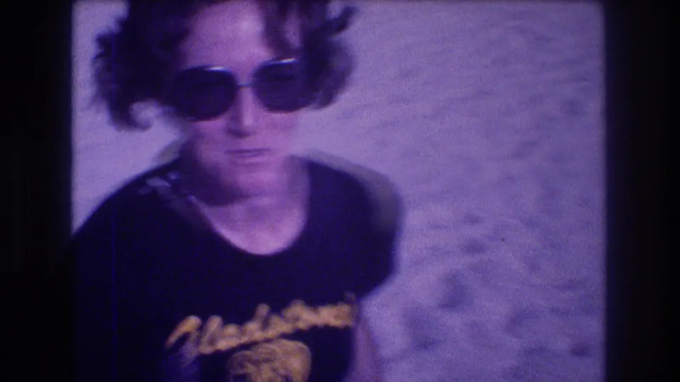 Close portrait of a woman in oversized dark sunglasses and black Gladstone's souvenir shirt on a sandy beach; metadata-dated Acapulco, Mexico, 1982, 8mm film with pronounced purple shift.