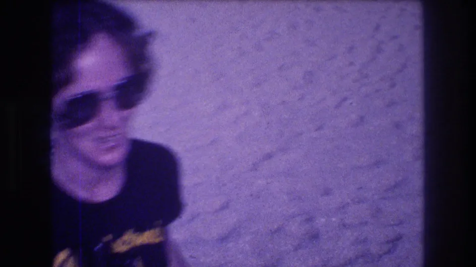 Motion-blurred frame of a woman turning on a sandy beach, metadata-dated Acapulco, Mexico, 1982; dark sunglasses and Gladstone's souvenir shirt partially visible through 8mm purple haze.