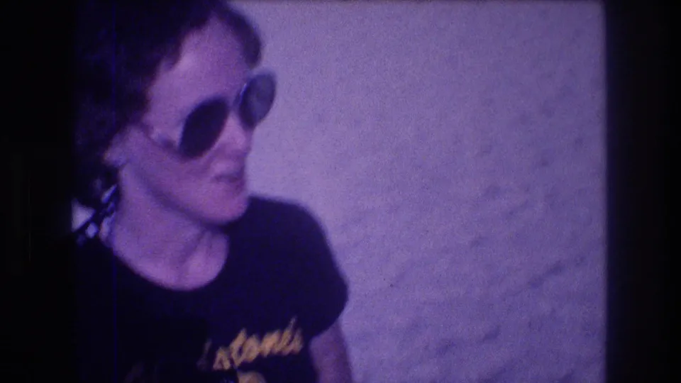 Woman in large dark sunglasses and black Gladstone's souvenir t-shirt on a sandy beach, metadata-dated Acapulco, Mexico, 1982, 8mm film with characteristic purple fading.