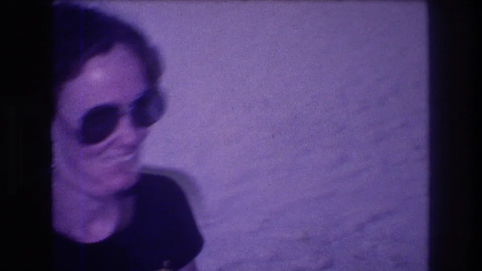 Cropped close-up of a woman in large 1980s-style sunglasses, from an 8mm home movie filmed in Acapulco, Mexico, circa 1982; pronounced purple color cast.