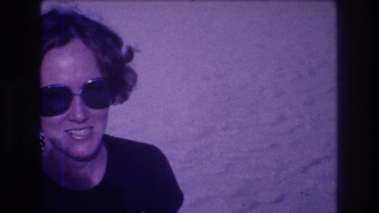 Woman with short curly hair and large dark sunglasses smiling, head and shoulders against a light textured wall; earring visible; purple-tinted film stock.