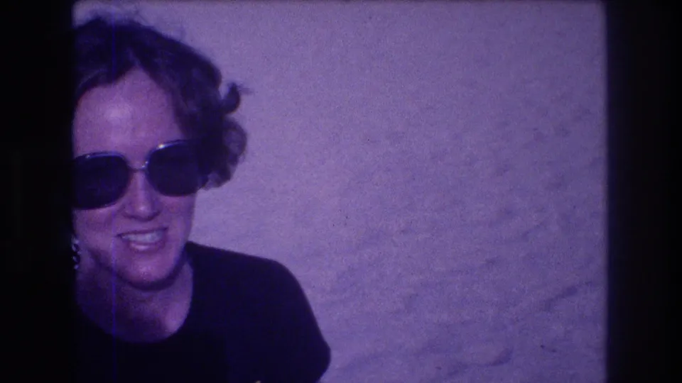 Woman in large-frame sunglasses smiling at the camera operator, head-and-shoulder shot from an 8mm vacation home movie, Acapulco, Mexico, 1982; purple film cast.