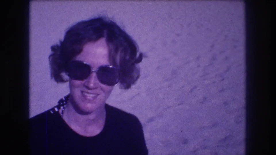 Woman in large-frame 1980s sunglasses and patterned necklace smiling broadly at the camera, from an 8mm home movie filmed in Acapulco, Mexico, 1982.