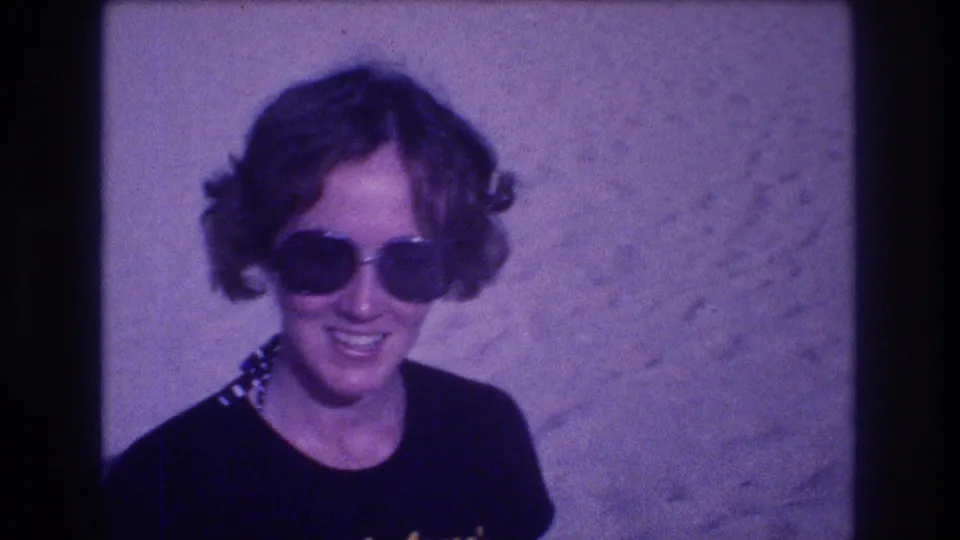 Woman with curly hair and oversized dark sunglasses smiling at the camera, likely Acapulco Mexico circa 1982, on 8mm film with strong purple color cast.
