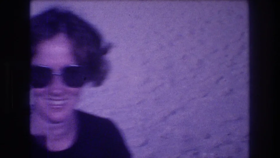 Woman with oversized dark sunglasses smiling as camera begins to pan away, motion blur visible, Acapulco Mexico circa 1982, 8mm film.