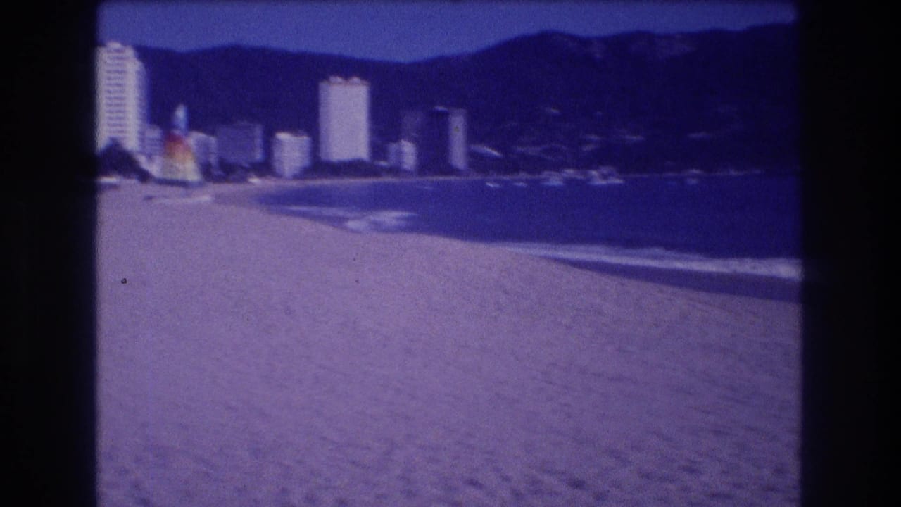 Sandy beach foreground with a row of white high-rise resort towers along the shoreline and green hills behind under a blue sky.