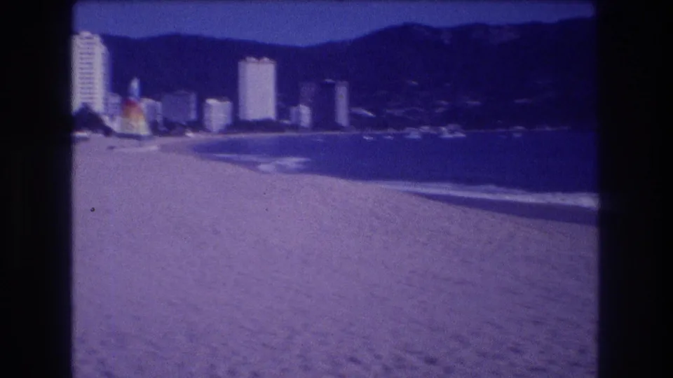 Wide beach pan showing Acapulco hotel-zone skyline circa 1982: white high-rise towers, green hillside, sandy beach, and blue-cast ocean water.
