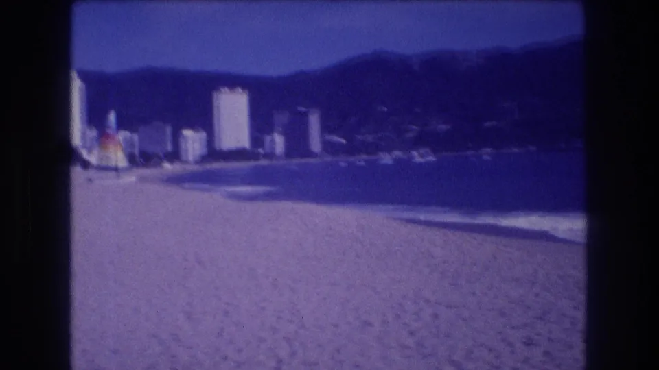 Acapulco beachfront circa 1982 showing high-rise hotels, sandy shore, a bright beach umbrella near the waterline, and forested hills in the background.