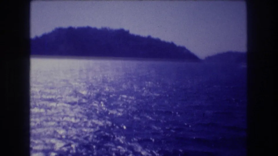 Ocean view toward a rocky forested headland in the Acapulco bay area circa 1982, with sun glitter on blue-purple water and visible film grain.