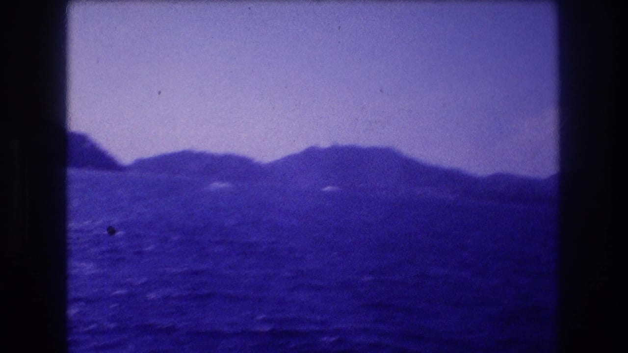 8mm home-movie frame c.1982 of a coastal mountain silhouette above dark blue water, likely Acapulco bay area, with aged blue-purple emulsion and a faint distant speck possibly indicating a vessel.