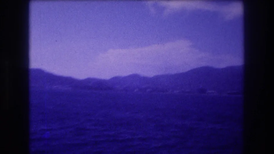 8mm home-movie frame c.1982 showing a wide pan of a coastal landscape with lavender sky, a haze-draped mountain range, and calm dark water, consistent with the Acapulco bay area.