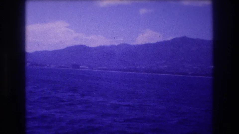 8mm home-movie frame c.1982 showing a wide coastal view with mountains, cloud wisps, dark blue water, and what may be small buildings along the distant shoreline, consistent with Acapulco bay.