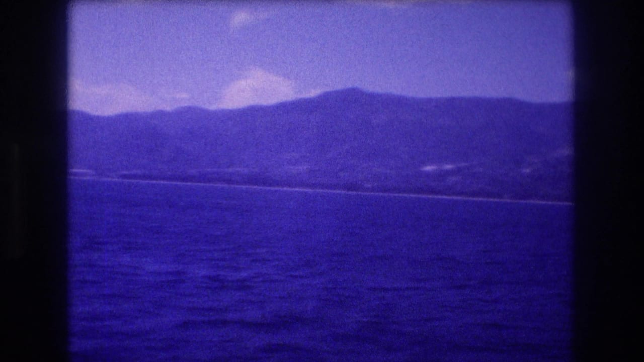 Open ocean view with distant mountain range and faint coastal development along the shoreline, heavy blue color cast throughout