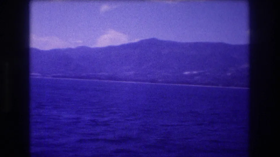 8mm home movie frame from Acapulco bay area, 1982 — wide ocean view with distant mountains and faint shoreline buildings under a blue-shifted sky