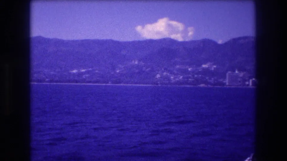 8mm home movie frame from Acapulco bay, 1982 — hotel zone skyline with backing mountain range, shot from a moving boat with heavy blue color cast