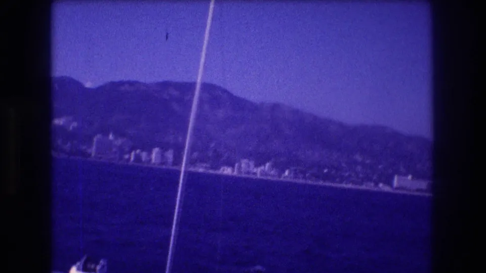 8mm home movie from Acapulco bay, 1982 — sailboat stay lines frame a wider view of the hotel strip and backing mountains under an open blue sky