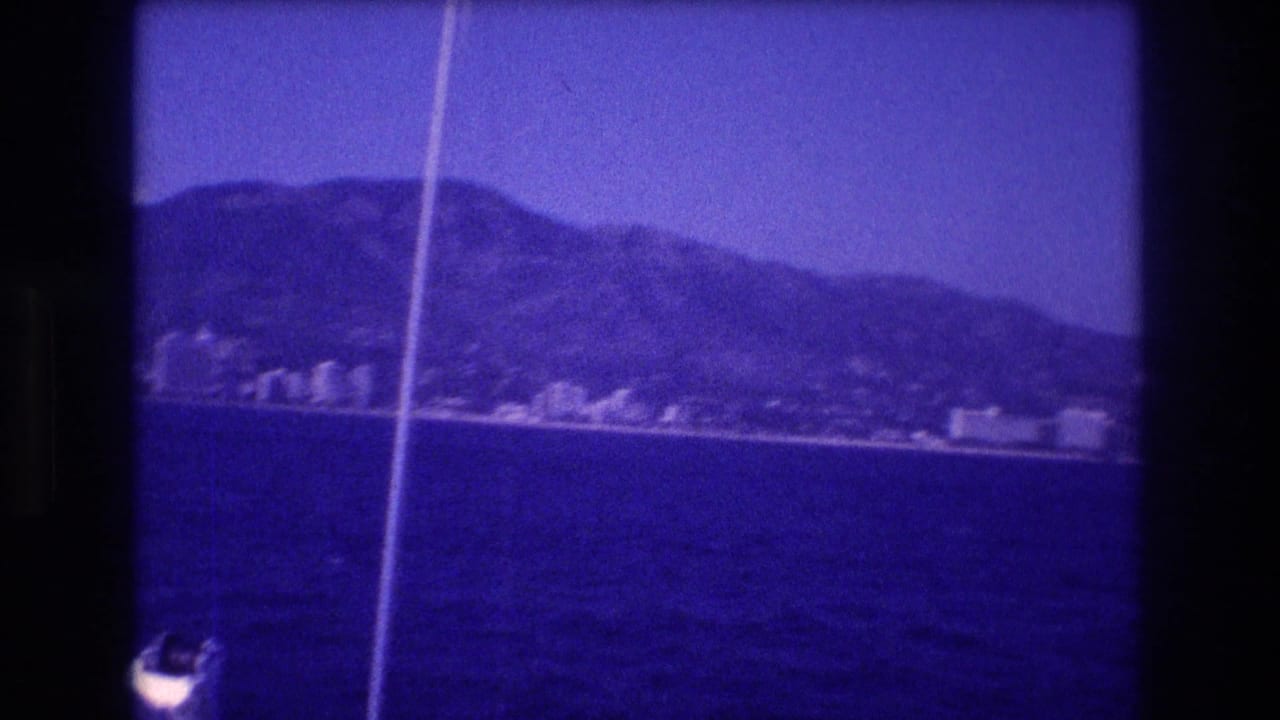 Panning boat shot of a mountainous bay coastline with low-rise buildings, heavy blue color cast, and a vertical white line artifact at the left edge.