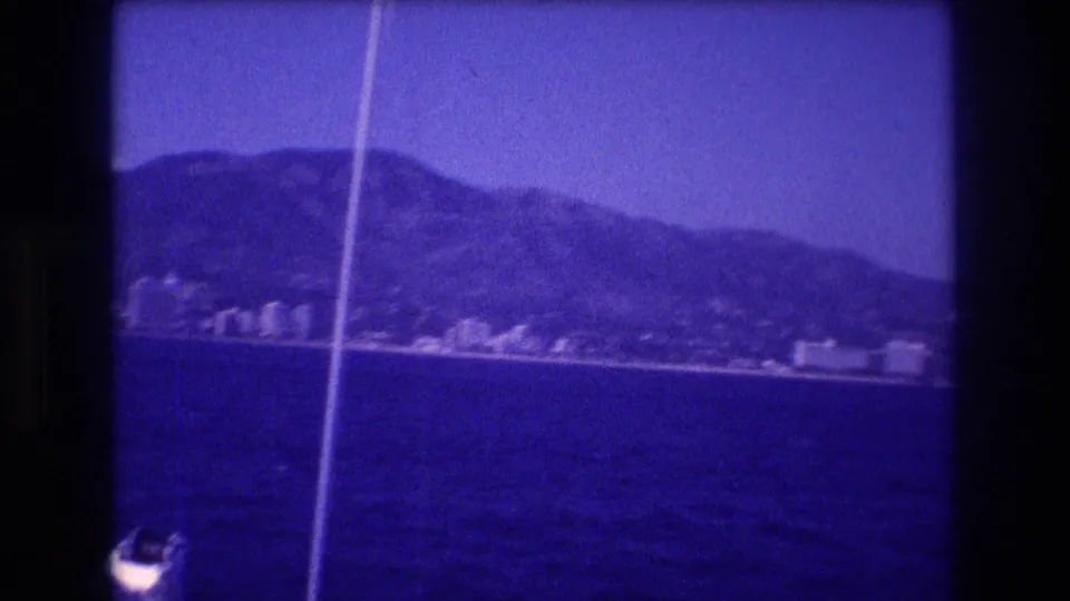 8mm home movie frame, circa 1982 Acapulco, Mexico: blue-shifted coastline with a steep ridge, white shoreline buildings, and a film scratch artifact at the left edge.