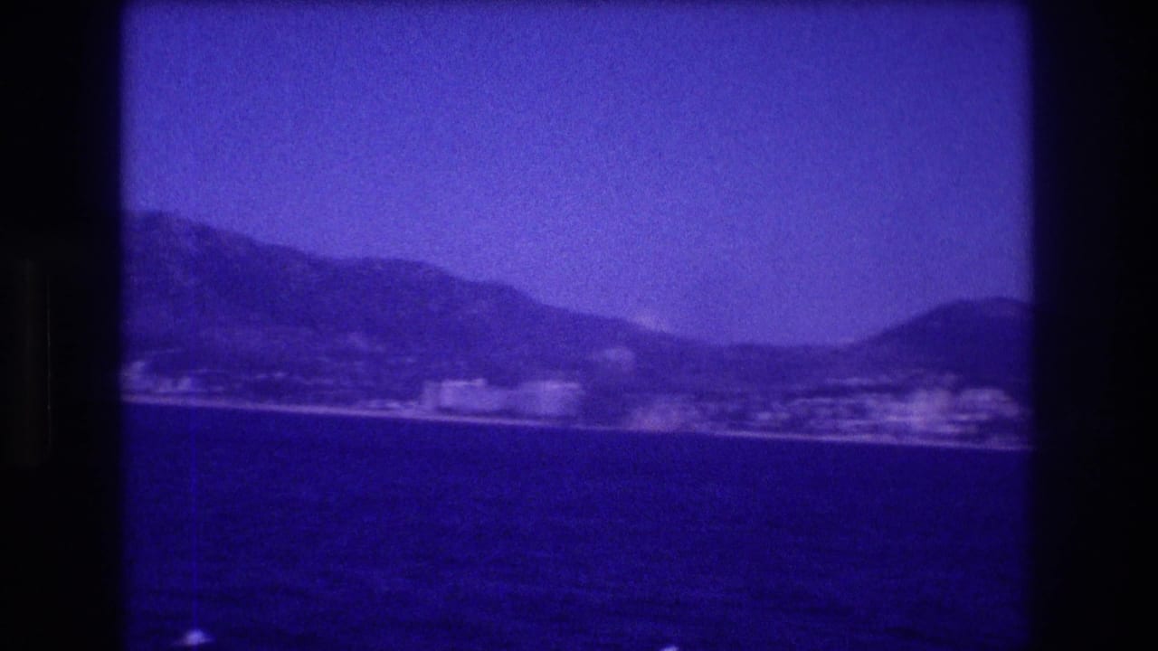 Panning view of a bay with a steep mountain ridge, clustered buildings along the shore, and a pronounced blue-violet film color cast.