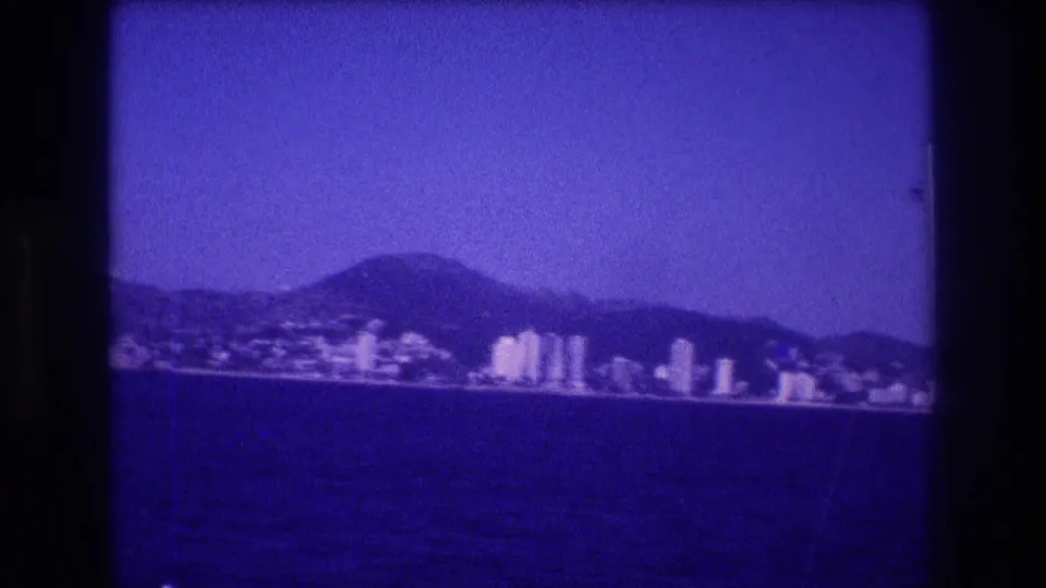 8mm home movie frame, circa 1982 Acapulco, Mexico: pan reveals a denser cluster of multi-story resort buildings along the waterfront, with lower hills behind and clear sky above.