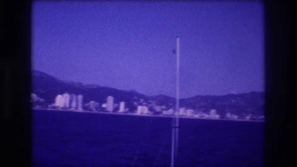 8mm home movie frame, circa 1982 Acapulco, Mexico: pan ends on a cluster of taller hotel towers at the waterfront, with a film scratch artifact visible at the right edge.