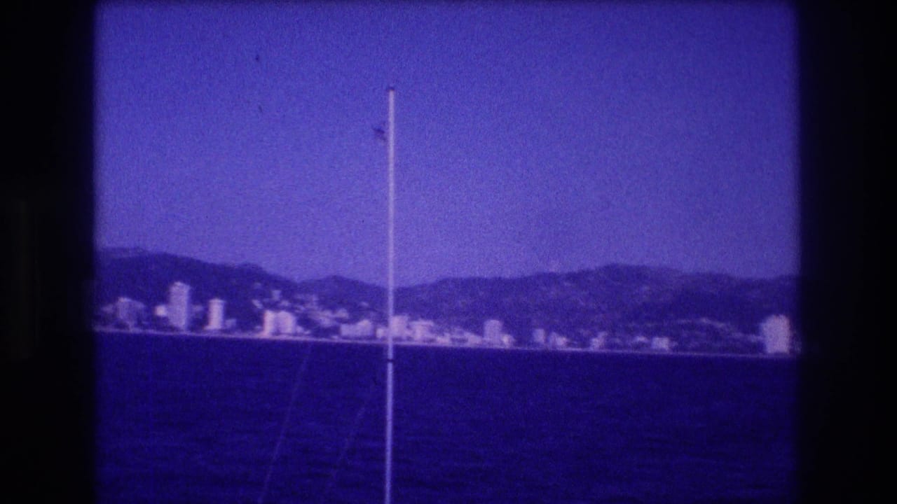 8mm home movie frame, circa 1982, showing Acapulco's resort skyline and mountain backdrop from a boat, blue dye-shift dominant