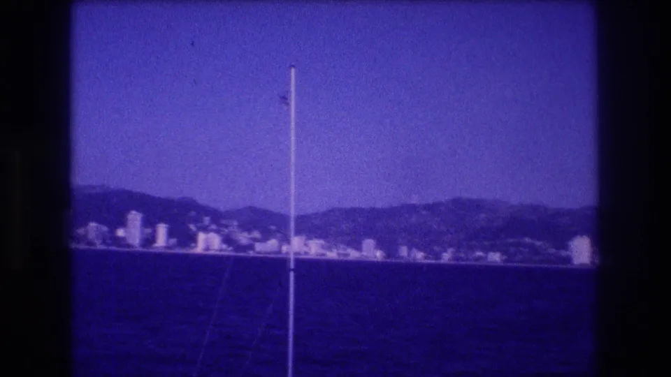 8mm home movie frame, circa 1982, showing Acapulco's resort skyline and mountain backdrop from a boat, blue dye-shift dominant