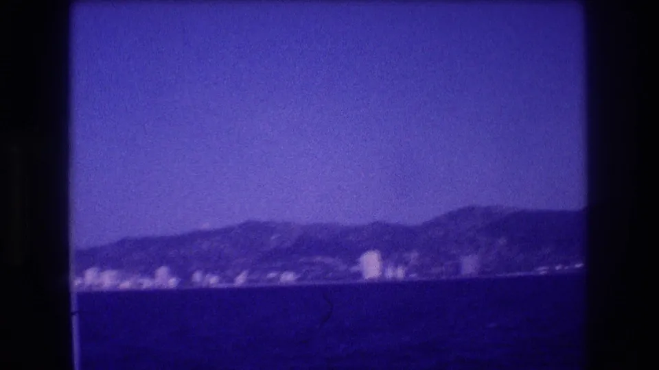 8mm home movie, circa 1982, wider boat-POV of Acapulco coastline showing mountains and distant resort strip under blue-shifted sky