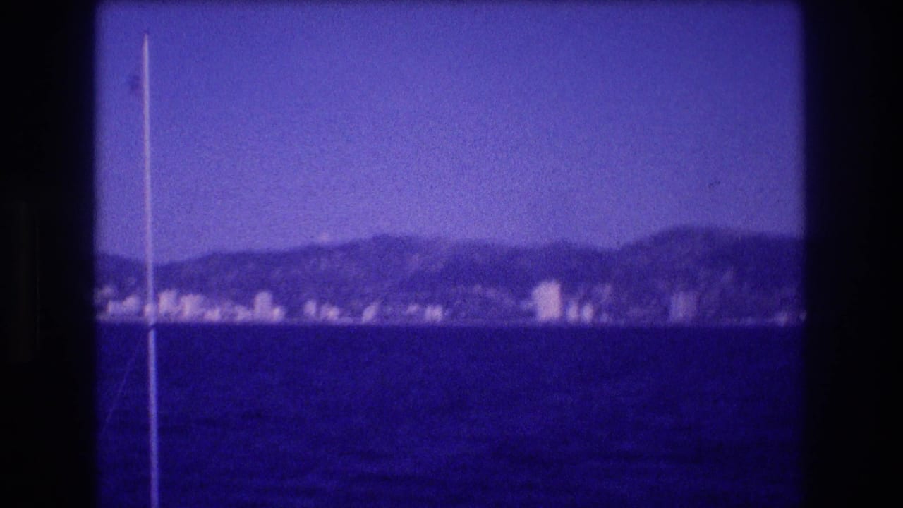8mm home movie frame, c. 1982, looking across calm blue-purple water toward what may be the Acapulco hotel zone, with a mast or flagpole at the left edge.