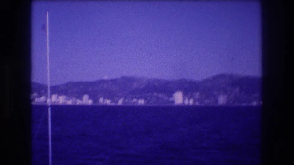 8mm home movie frame, c. 1982, looking across calm blue-purple water toward what may be the Acapulco hotel zone, with a mast or flagpole at the left edge.