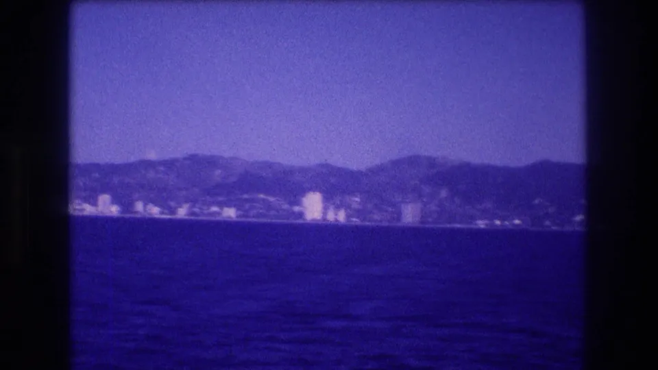 8mm home movie frame, c. 1982, showing a blue-purple bay and a distant cluster of hotel-style towers beneath a hilly ridge, likely the Acapulco waterfront.