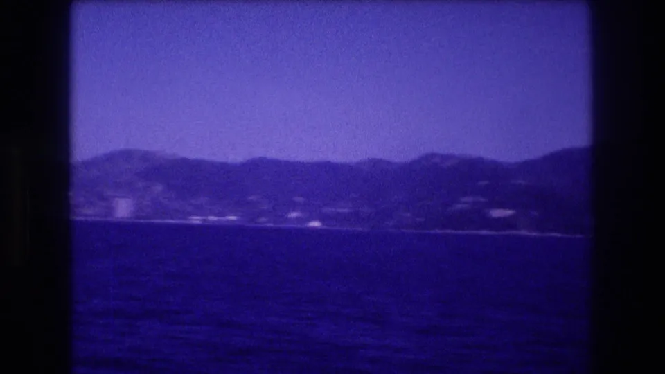 8mm home movie frame, c. 1982, showing a pan or drift that brings a hillside silhouette into frame as the built-up shoreline recedes to the left.