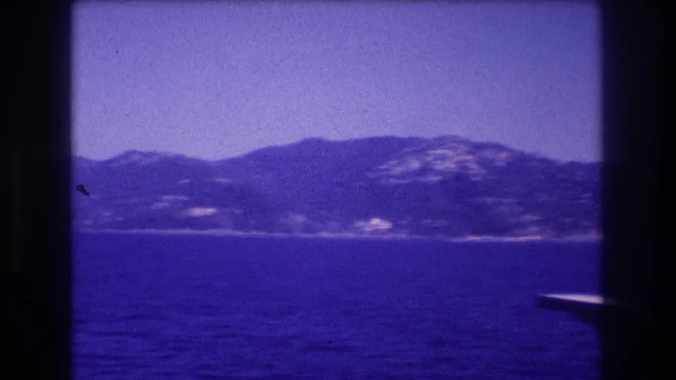8mm home movie frame, c. 1982, with a hilly coastal silhouette and the corner of a boat structure entering the lower-right frame edge.