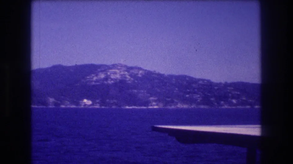 Blue-tinted 8mm frame from the supplied 1982 Acapulco footage showing open water, a hazy mountain shoreline, and the edge of a boat in the foreground.
